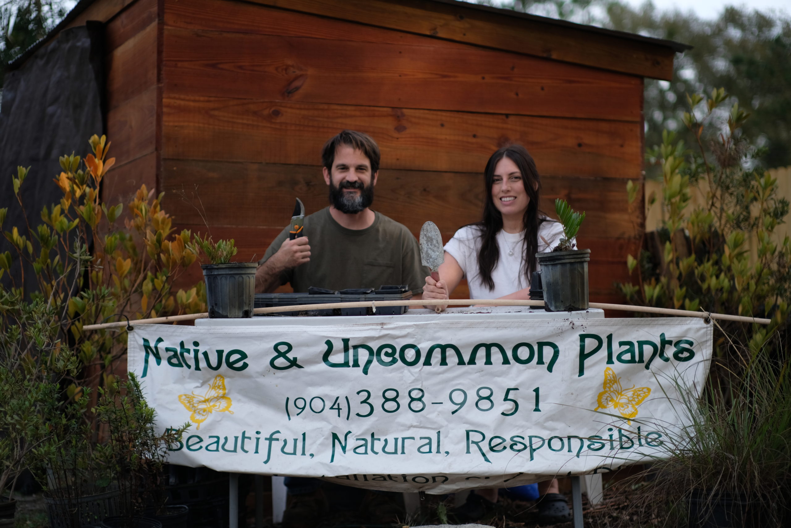 Alfred and Leslie sitting behind a table with a sign in the front that has Native & Uncommon Plants with the phone number and 3 adjectives. Leslie is wearing white sitting on the right while Afred is wearing dark green sitting on the left. Both are smiling proudly holding up gardening tools